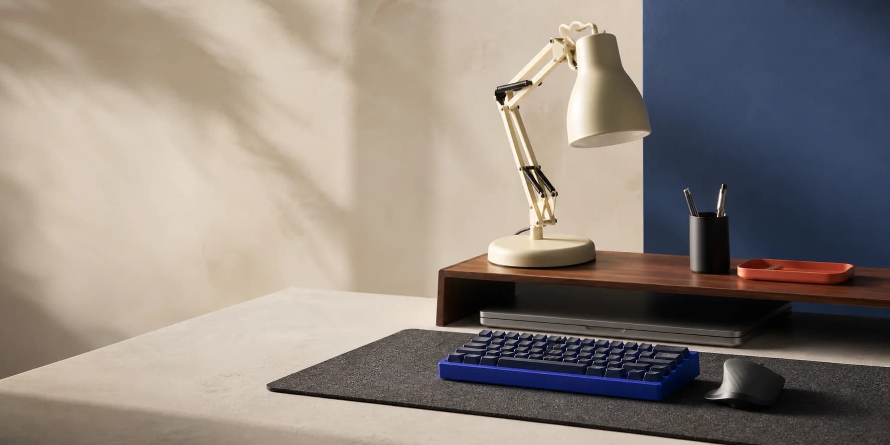 A considered workspace with a cobalt keyboard, ivory lamp, walnut riser, and graphite desk mat